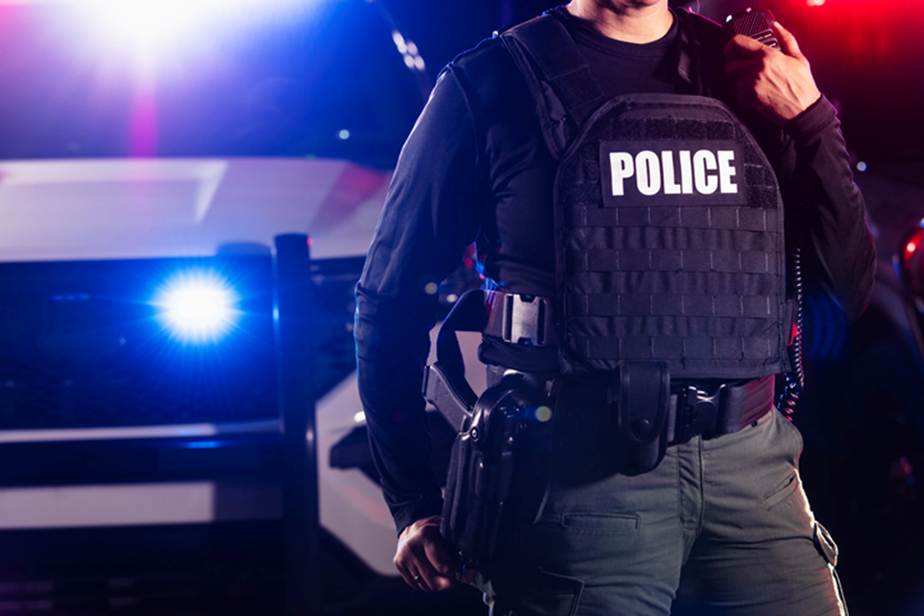 A police officer stands at night near a vehicle with flashing lights. The officer wears a vest marked "POLICE," and carries a radio.